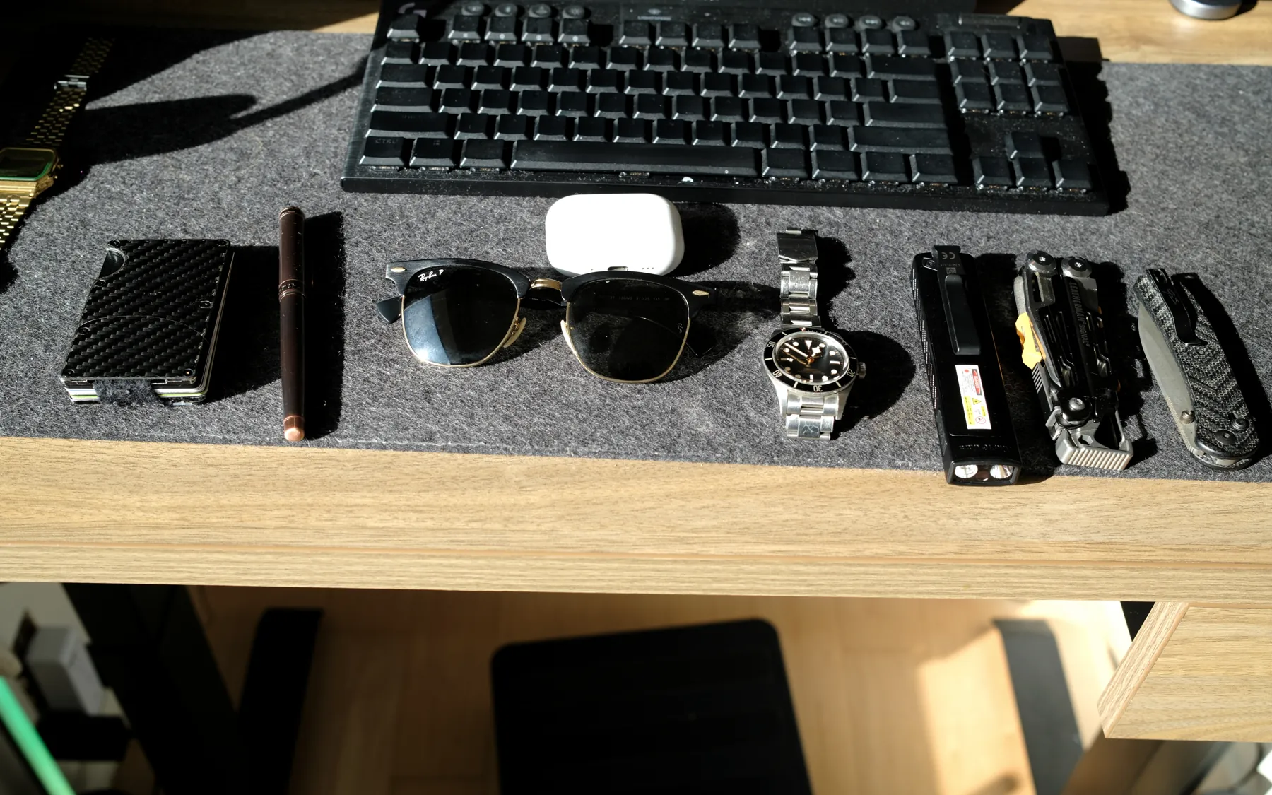 Top-down photo of a wallet, pens, sunglasses, earbuds, two watches, a flashlight, a multi-tool and a knife on a felt desk mat